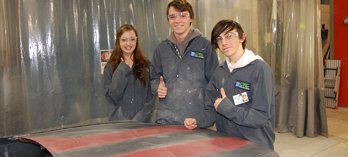 Two boys and a girl standing in automotive lab showing the hood of a car that is being repaired and painted.